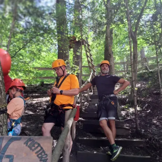 Eine Gruppe von Menschen im Wald, mit Helmen und Gurten. Ein Mann sitzt auf einer Holzschiene, ein anderer steht auf einer Treppe. Dahinter eine Holzumzäunung und ein Baum mit einem roten Ballon.