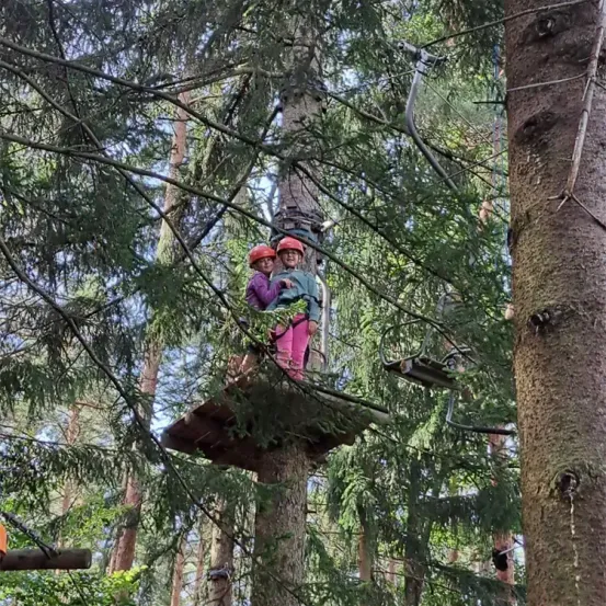 Zwei junge Kinder in Helmen stehen zusammen auf einer Plattform in einem Baum, umgeben von dichtem Wald. Im Hintergrund ist ein Seilbahnsystem zu sehen.