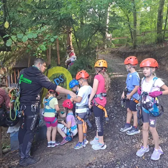 Eine Gruppe von Kindern in Helmen steht draußen. Ein Kind sitzt auf dem Boden. Ein Mann unterrichtet sie. Dahinter steht ein gelbes Objekt mit dem Wort 'DS' darauf. Es gibt Bäume und Pflanzen um sie herum.