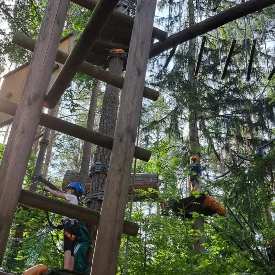 Zwei Kinder sind in einem Wald und klettern eine Holzstruktur, tragen Helme und Gurte. Ein Kind klettert, während das andere auf einer Plattform ist.