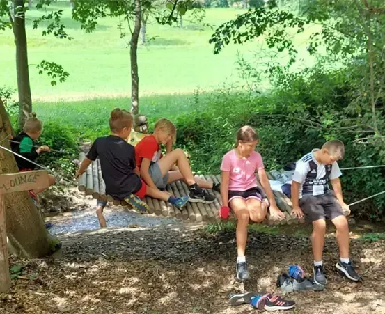 Kinder sitzen auf einer hölzernen Hängebrücke im Wald, tragen Freizeitkleidung. Schuhe sind darunter abgestellt, und ein Holzschild steht 'CHILER LOUNGE'. Bäume und Büsche umgeben das Areal.