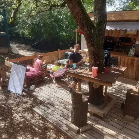Ein Mann und zwei Kinder sitzen an einem Holztisch unter einem Baum auf einer Holzterrasse. Eine Frau ist in der Nähe auf einer Bank. Im Hintergrund hat ein kleines Gebäude einen Tisch und Stühle.
