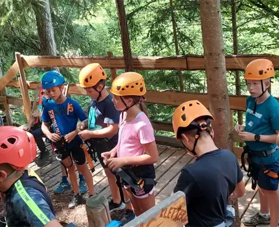 Eine Gruppe von Kindern, alle mit Schutzhelmen, steht auf einer Holzplattform in einem Waldgebiet. Einige von ihnen halten ihre Gurte.