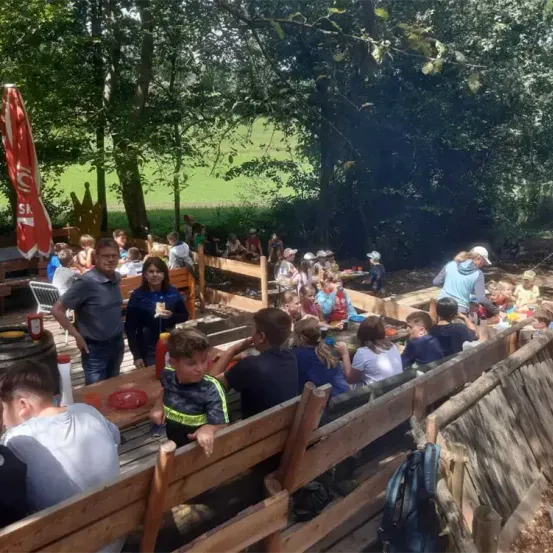 Eine Gruppe von Menschen, einschließlich Erwachsener und Kinder, essen im Freien an einem hölzernen Picknicktisch unter einem roten Schirm. Bäume und ein grünes Feld sind im Hintergrund zu sehen.