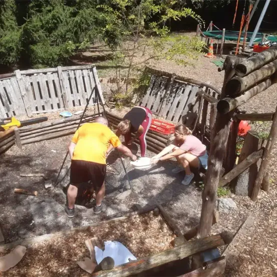 Drei Personen arbeiten an einer Holzstruktur in einem Außenbereich. Ein Mann beugt sich vor, während zwei Frauen ein weißes Objekt halten. Der Bereich ist von Holzzäunen umgeben.
