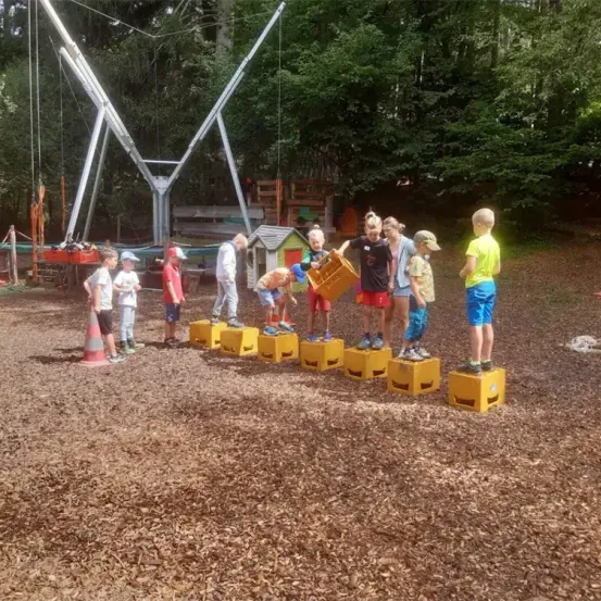 Mehrere Kinder stehen auf gelben Kisten in einem Park. Ein Junge trägt einen Korb, während ein anderer einen Helm trägt. Im Hintergrund befinden sich eine Schaukel und ein Holzhaus.