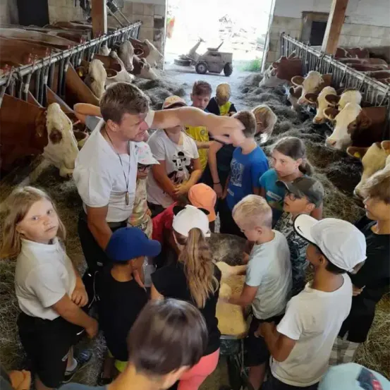 Ein Mann zeigt einer Gruppe von Kindern, wie man eine Kuh füttert. Die Kinder tragen Mützen und Turnschuhe. Es gibt Kühe im Stall.
