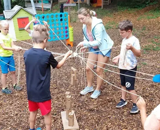 Eine Gruppe von Kindern spielt auf einem Spielplatz, wobei ein Junge eine mit einer Struktur verbundene Schnur hält. Eine Frau steht in der Nähe.