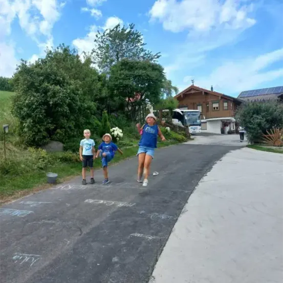 Drei Kinder spielen Hüpfspiel auf einer Einfahrt. Ein Kind springt, während die anderen beiden zuschauen. Ein Haus ist im Hintergrund.