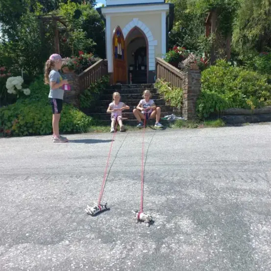 Drei Kinder spielen mit einem Spielzeugauto auf einer Straße, mit einer kleinen Kapelle im Hintergrund.