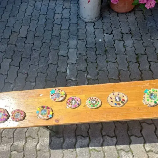 Eine Holzbank mit mehreren bunten kreisförmigen Objekten darauf. Die Objekte befinden sich auf einer Kopfsteinpflasteroberfläche. Im Hintergrund steht ein Topf mit rosa Blumen.