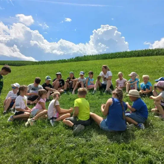 Eine Gruppe von Kindern und einer Frau sitzt im Kreis auf einer Grasfläche unter einem bewölkten Himmel. Die Frau steht und scheint mit den Kindern zu sprechen. Die Kinder tragen Mützen und Turnschuhe. Die Frau trägt eine Uhr und Turnschuhe. Die Kinder schauen auf etwas am Boden.