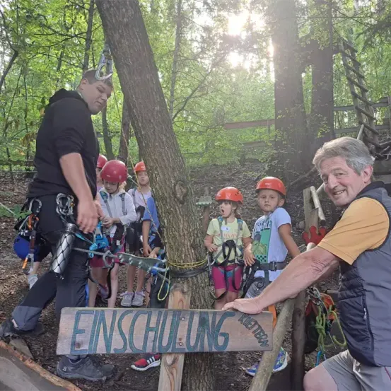 Eine Gruppe von Kindern mit Helmen im Wald. Ein Mann gibt Anweisungen. Es gibt ein Holzschild mit der Aufschrift Einschulung.