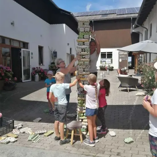 Eine Gruppe von Kindern und Erwachsenen baut einen Stapel aus Blöcken in einem Außenbereich. Sie stehen um einen hohen Stapel herum, wobei eine Frau nach oben greift.