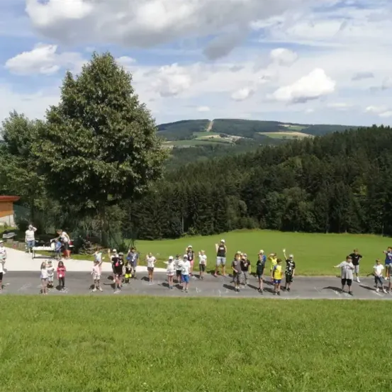 Eine Gruppe von Menschen, möglicherweise eine Schule, steht in einer Reihe auf einem Pfad in einem Grasfeld. Dahinter befinden sich Bäume und eine entfernte Aussicht auf Berge.