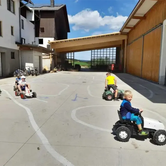 Vier Kinder fahren mit Spielzeugautos auf einem Parkplatz. Hinter ihnen steht ein Gebäude mit einem Vordach. In der Ferne gibt es ein Feld und einen Berg.