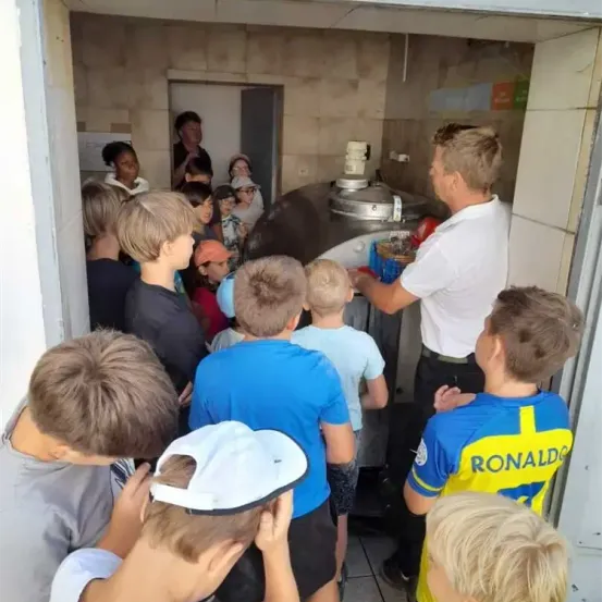 Eine Gruppe von Kindern ist in einer Küche um einen Mann versammelt und beobachtet ihn interessiert. Er trägt ein Shirt mit dem Namen 'Ronaldo'.