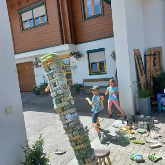 Zwei Kinder spielen mit einem Eierkartonturm vor einem Haus. Ein Junge hält einen Stock, während ein Mädchen läuft. Eine Bank und Pflanzen sind im Hintergrund.