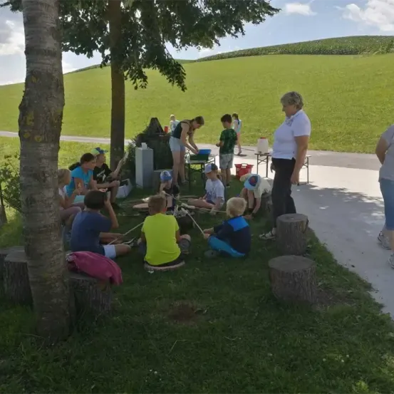 Kinder sitzen auf einer Grasfläche um einen Baumstumpf, eine Frau steht in der Nähe. Einige sind mit Aktivitäten beschäftigt, während andere beobachten. Eine Frau in der Ferne beugt sich über etwas. Tische mit Gegenständen und Eimern sind aufgestellt. In der Ferne ein grünes Feld und Hügel unter einem blauen Himmel.
