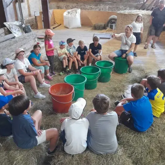 Eine Gruppe von Kindern sitzt im Kreis auf Heu und hört einer Frau zu, die spricht. Grüne und orangefarbene Eimer sind um sie herum platziert. Im Hintergrund stehen zwei Erwachsene.