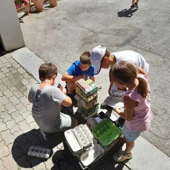 Vier Kinder sitzen auf dem Bürgersteig und sortieren und stapeln Eierkartons. Ein Kind kniet, ein anderes steht, und die anderen sitzen. Tabletts und Körbe mit Eiern sind um sie herum.