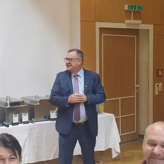 Ein Mann in Anzug und Krawatte steht lächelnd vor einem Tisch, mit einer Tür und einem Buffet dahinter.