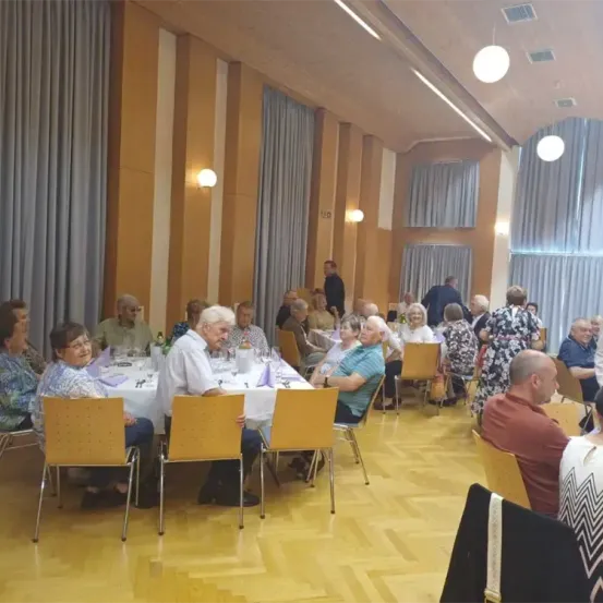 Ein Speisesaal mit mehreren älteren Menschen, die an Tischen mit weißen Tischdecken sitzen. Der Saal hat Holzböden, gelbe Wände und mehrere Lampen. Ein paar Leute stehen in der Nähe der vorhangbehangenen Wand.