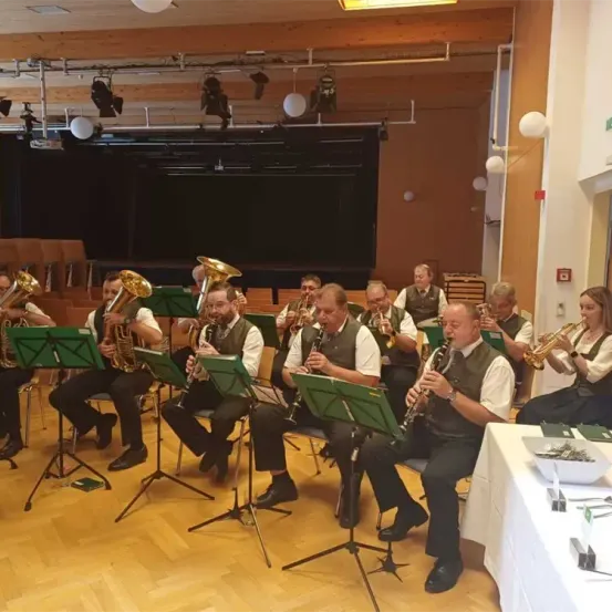 Eine Gruppe von Musikern sitzt in einem Raum und spielt ihre Instrumente. Der Raum ist gut beleuchtet. Es gibt Stühle, Tische und Notenständer im Raum.