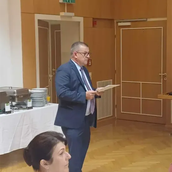 Ein Mann in einem Anzug steht in einem Raum und liest von einem Blatt Papier. Eine Frau hört ihm zu. Hinter ihm steht ein Tisch mit Tellern und Essen.