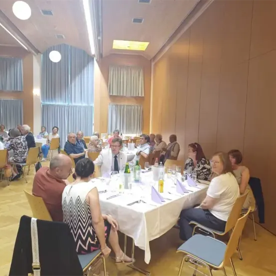 Eine Gruppe von Menschen sitzt um einen langen Tisch in einem Speisesaal und unterhält sich. Flaschen, Gläser und andere Gegenstände stehen auf dem Tisch. Der Raum hat Holzwände und Vorhänge.