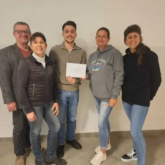 Fünf Personen stehen lächelnd in einem Raum. Ein Mann in der Mitte hält eine Urkunde. Die beiden Frauen tragen Brillen und Turnschuhe, und die Männer tragen Brillen und Stiefel.