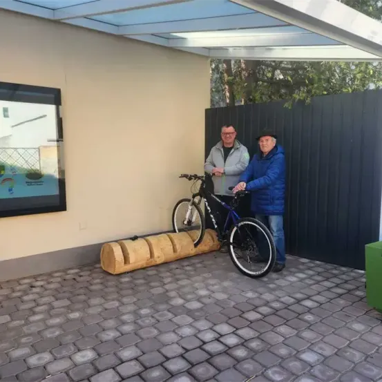 Zwei Männer stehen neben einem Fahrrad in einem überdachten Bereich. Ein Mann hält das Fahrrad, während der andere einen Holzstamm hält. Ein Bilderrahmen ist an der Wand montiert.
