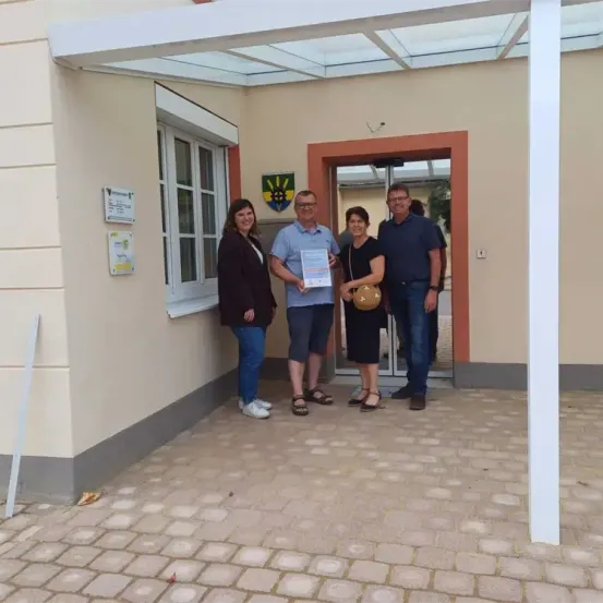 Vier Personen, zwei Frauen und zwei Männer, stehen vor einem Gebäude, einer hält eine Urkunde. Sie sind lässig gekleidet. Hinter ihnen ist eine Glastür mit rotem Rahmen und Wappenschild zu sehen.