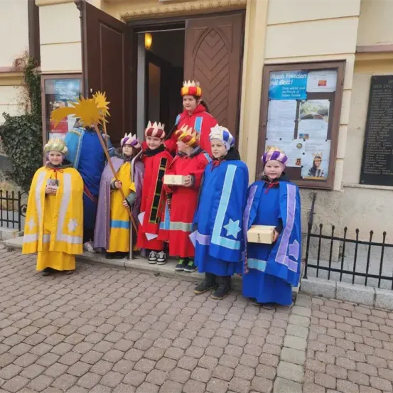 Eine Gruppe von Kindern in farbenfrohen Kostümen mit Kronen steht vor einem Gebäude. Sie halten eine Box und scheinen für ein Foto zu posieren.