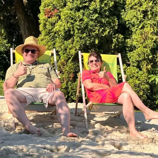 Zwei Personen sitzen auf Strandstühlen an einem Sandstrand und geben beide Daumen hoch, mit einem grünen Busch im Hintergrund.