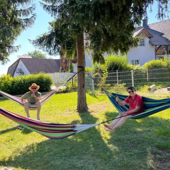 Zwei Menschen entspannen in Hängematten im Hinterhof mit Häusern und Bäumen im Hintergrund.