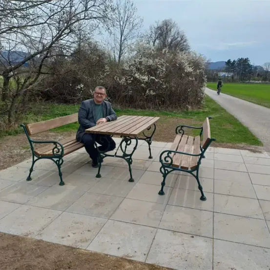 Ein Mann sitzt auf einer Parkbank mit Tisch, in der Nähe eines grünen Feldes.