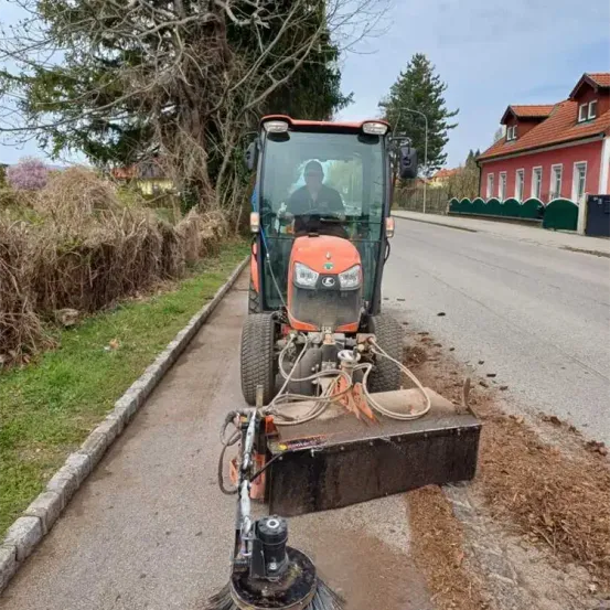 Ein Traktor mit einem großen Bürstenaufsatz räumt Blätter von der Straße, mit einem roten Gebäude und einem grünen Zaun im Hintergrund.