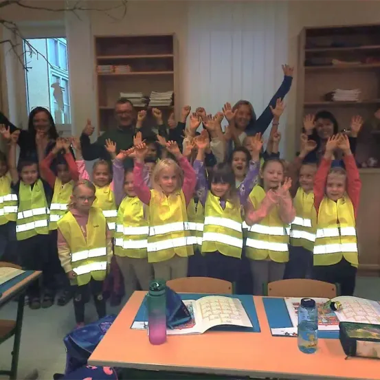 Ein Klassenzimmer mit Kindern in gelben Westen, die die Hände heben, ein Lehrer mit Brille und ein Buch auf einem Tisch im Vordergrund.