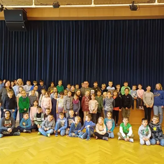 Eine Gruppe von Kindern und Erwachsenen posiert für ein Foto in einem Raum mit Holzboden und einer Bühne mit blauen Vorhängen.