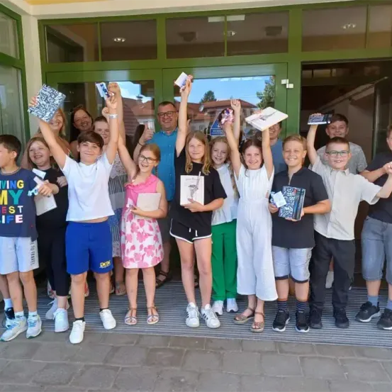 Eine Gruppe von Kindern und Erwachsenen posiert für ein Foto vor einem Gebäude, hält Bücher hoch und lächelt.