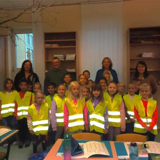 Eine Gruppe von Kindern und Erwachsenen steht in einem Klassenzimmer. Die Kinder tragen reflektierende Westen. Die Erwachsenen, darunter zwei Frauen, stehen hinter den Kindern. Der Raum hat Bücherregale, ein Fenster und einen Tisch mit Büchern.