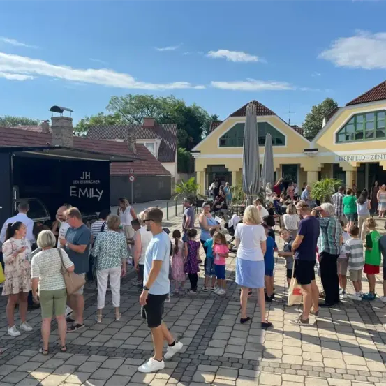 An einem sonnigen Tag versammeln sich Menschen um einen Food-Truck und Gebäude mit Namen wie 'JH der Eismacher Emily' und 'Steinfeld Zent'. Einige warten in der Schlange, andere unterhalten sich, und es gibt spielende Kinder.