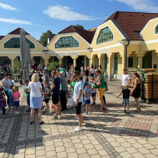 Eine Gruppe von Menschen steht vor einem gelben Gebäude mit braunen Dächern, einige tragen Hüte und Sonnenbrillen. Ein Stapel Holzpaletten steht rechts.