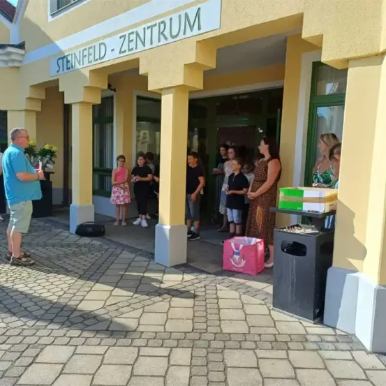 Eine Gruppe von Menschen, einschließlich Kindern, steht vor dem Steinfeld Zentrum, einem gelben Gebäude mit Glasfenstern. Ein Mann hält einen Blumenstrauß, und ein Mülleimer mit einem rosa Beutel befindet sich in der Nähe.