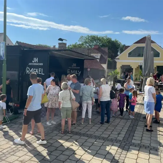 Eine Gruppe von Menschen versammelt sich vor einem Food Truck. Sie stehen und unterhalten sich. Der Truck ist mit JH Eismacher und Rafael Emily beschriftet.