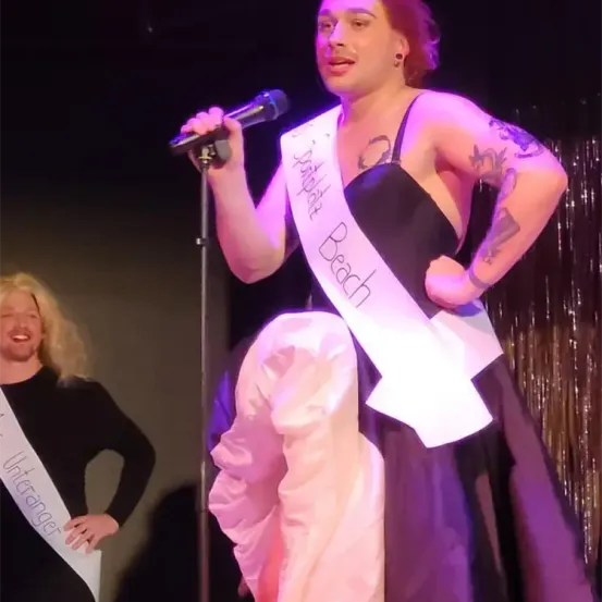 Eine Frau mit Tätowierungen an den Armen und einer Schärpe mit der Aufschrift 'Miss Pirate Beach' singt in ein Mikrofon. Ein Mann mit einer Schärpe, die 'Mr. Utrange' liest, steht hinter ihr und lächelt.