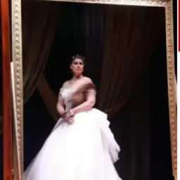 Eine Frau in einem weißen Kleid steht vor einem Spiegel mit goldenem Rahmen. Sie lächelt und hält einen Armreif in ihrer rechten Hand.