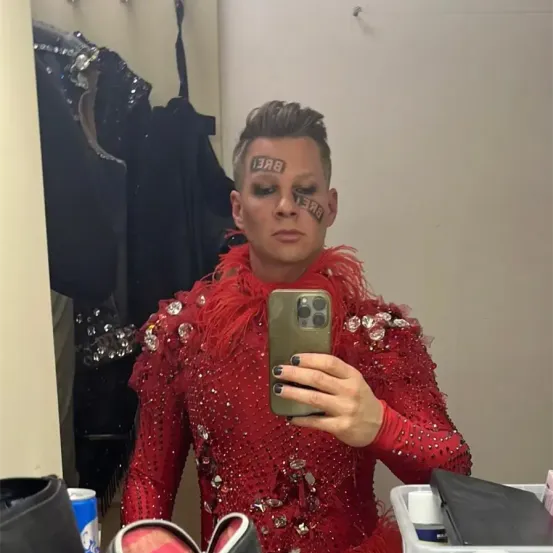 Eine Person in einem roten Paillettenkostüm mit federigen Details macht ein Selfie. Sie trägt schwarzen Eyeliner und Make-up im Gesicht, einschließlich des Wortes 'BABE' auf der Stirn.