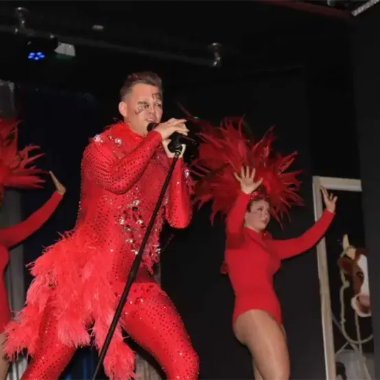 Ein Mann tritt auf der Bühne auf, begleitet von zwei tanzenden Frauen, alle in roten Pailletten-Outfits. Der Mann hält ein Mikrofon und die Frauen heben ihre Hände. Die Bühne ist schwach beleuchtet.